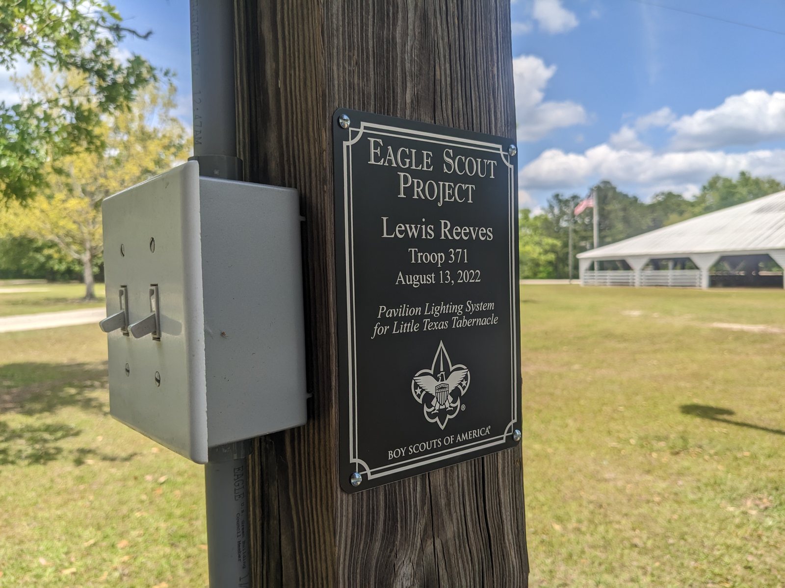 An Eagle Scout project plaque mounted on a pavilion post beside a two-gang weatherproof switch box and PVC conduit, with the pavilion visible behind.