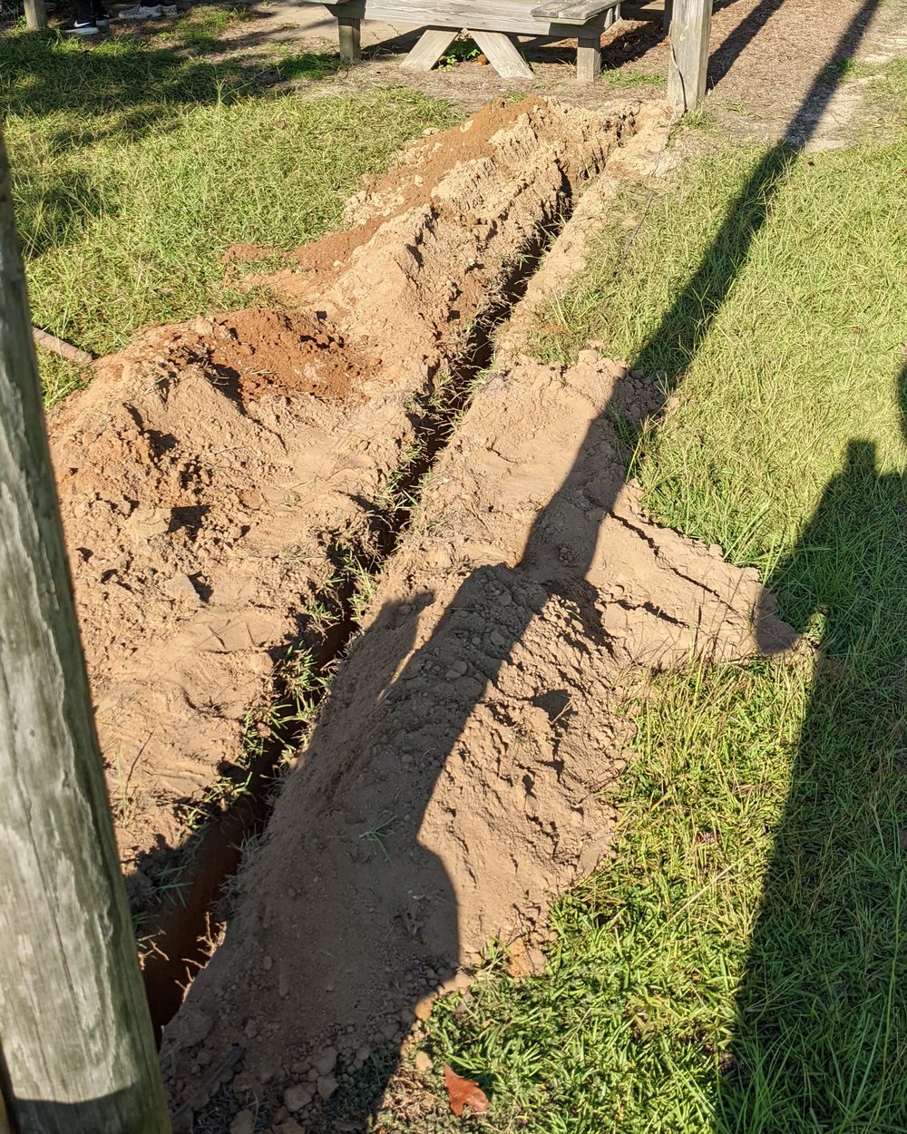 An open trench roughly eighteen inches deep running across grass between pavilion support poles.